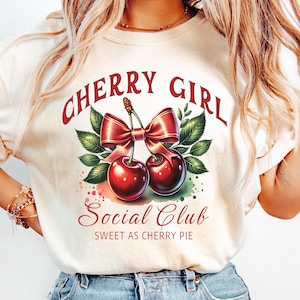 May include: A white t-shirt with a graphic design of two red cherries with a red bow and green leaves. The text "Cherry Girl Social Club Sweet as Cherry Pie" is printed on the shirt.
