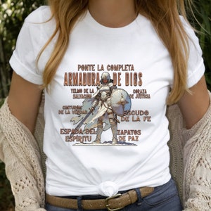 Pode incluir: Camiseta branca com um gr&aacute;fico de uma mulher com armadura, com o texto "PONTE LA COMPLETA ARMADURA DE DIOS" e uma lista de pe&ccedil;as de armadura em espanhol.