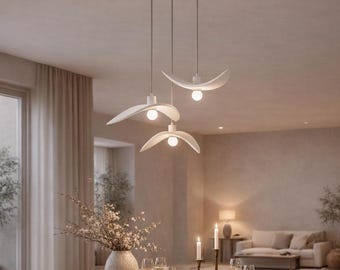 May include: A set of three white pendant lights with curved, wing-like shades suspended at varying heights. Each fixture features a single downward-facing bulb, creating a soft glow above a wooden dining table set with plates and candles.