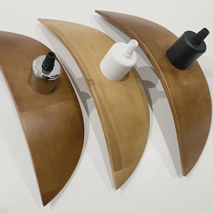 May include: Three wooden, crescent-shaped light fixtures in varying shades of brown and tan. Each fixture holds a different colored light socket: silver, white, and black. The fixtures are arranged on a white surface.