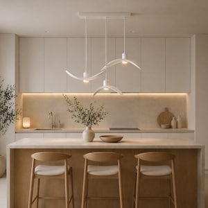 May include: A modern kitchen with a light color palette. Features include a central island with three wooden stools, a white pendant light fixture, and a neutral-toned countertop. A vase of greenery and a potted plant add natural elements.