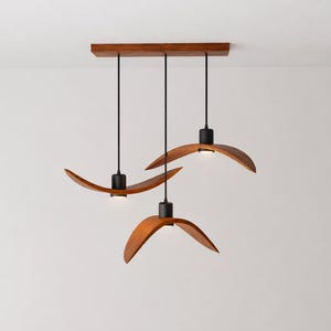 May include: A modern, three-light pendant lamp with a rectangular wooden canopy. Each light features a curved, wooden shade with a black fixture and a small, integrated LED light. The design is minimalist and elegant, suitable for various interior styles.