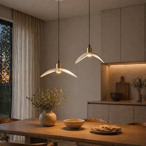 May include: Two modern pendant lights with a white, curved shade and gold accents hang above a wooden dining table. The room features a neutral color palette with a vase of greenery and a wooden bowl.