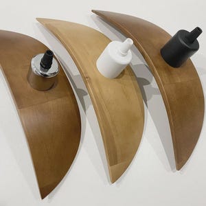 May include: Three wooden, crescent-shaped light fixtures in varying shades of brown and tan. Each fixture holds a different colored light socket: silver, white, and black. The fixtures are arranged on a white surface.