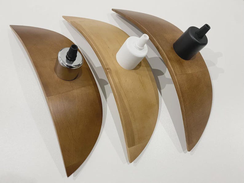 May include: Three wooden, crescent-shaped light fixtures in varying shades of brown and tan. Each fixture holds a different colored light socket: silver, white, and black. The fixtures are arranged on a white surface.