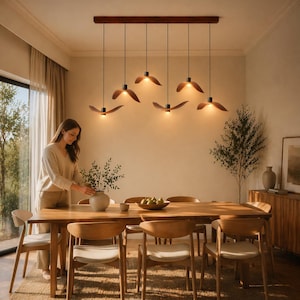 May include: A dining room scene featuring a wooden table and chairs, with a woman arranging a vase of greenery. Above the table, six pendant lights with wooden leaf-shaped shades hang from a dark brown beam.