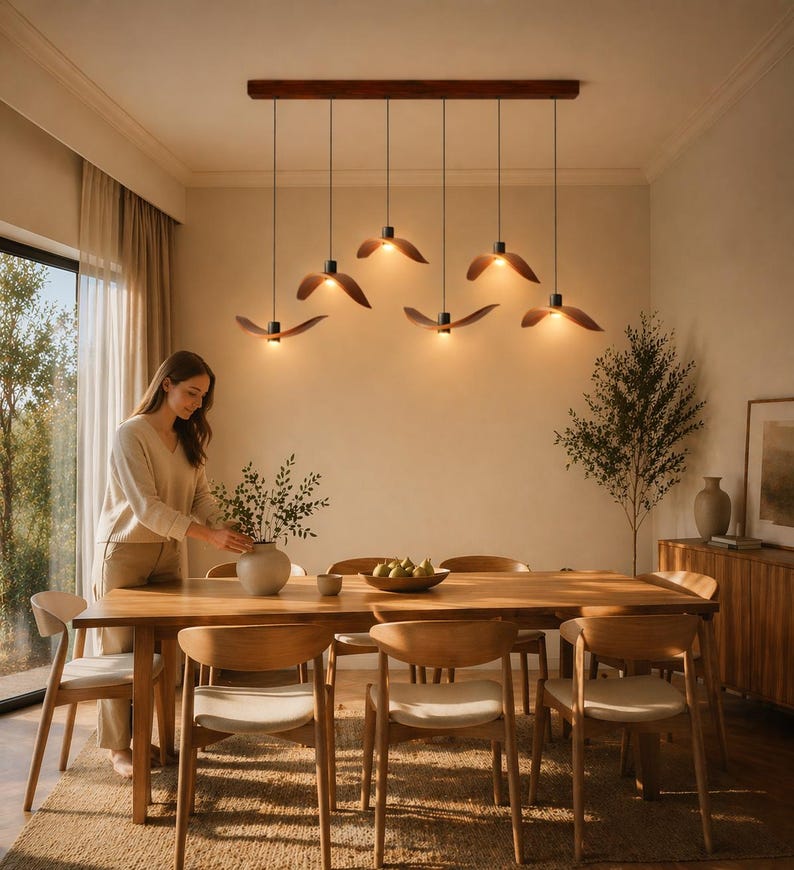 May include: A dining room scene featuring a wooden table and chairs, with a woman arranging a vase of greenery. Above the table, six pendant lights with wooden leaf-shaped shades hang from a dark brown beam.