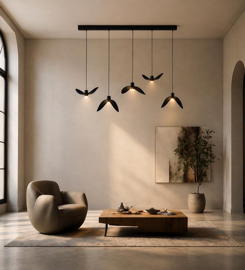 May include: Modern black pendant lights with a unique, leaf-like design hang from a black rectangular fixture. The lights are suspended above a living room with a neutral color palette, a brown armchair, and a wooden coffee table.