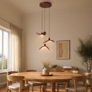 May include: A modern dining room featuring a wooden oval table and matching chairs. Three pendant lights with wooden shades hang above the table. The room has a neutral color palette with a large window and a sideboard.