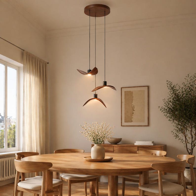 May include: A modern dining room featuring a wooden oval table and matching chairs. Three pendant lights with wooden shades hang above the table. The room has a neutral color palette with a large window and a sideboard.