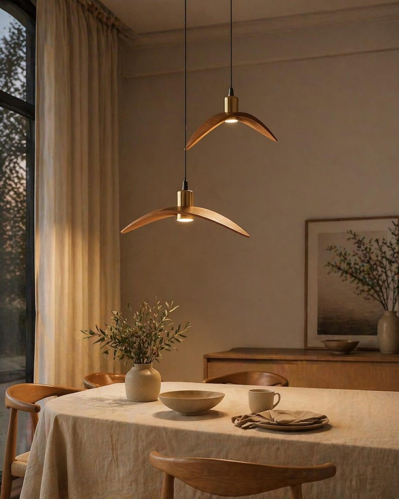 May include: Two modern wooden pendant lights with brass accents hang above a dining table. The lights have a curved, wing-like design. The table is set with a beige tablecloth, a bowl, a cup, and a vase with greenery, creating a warm and inviting atmosphere.