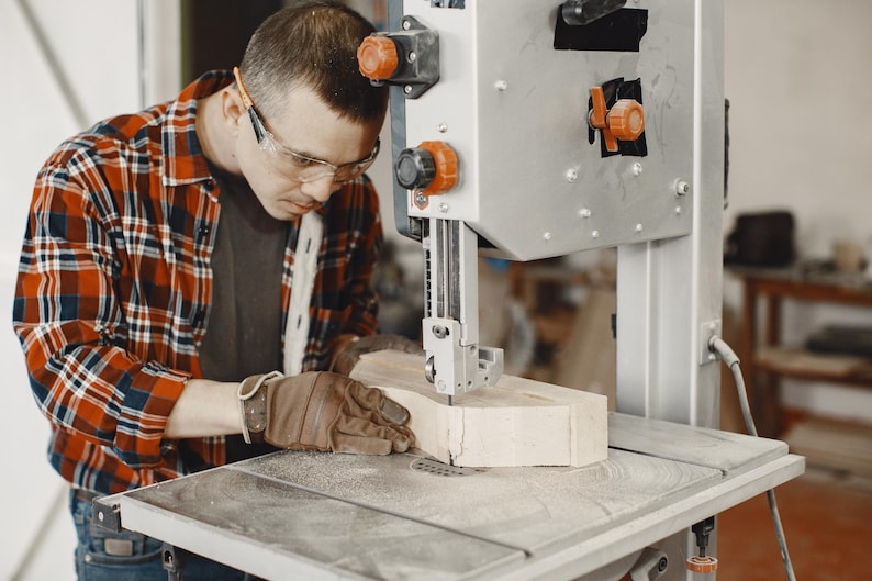 May include: A person wearing safety glasses and gloves operates a band saw, cutting a piece of wood. The saw is silver with orange accents. The person is wearing a red, black, and white plaid shirt.