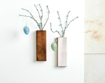 May include: Two wooden wall vases with a light brown and white finish, each holding a sprig of branches with blue blossoms and a decorative Easter egg hanging from the branch. The egg in the brown vase is blue and the egg in the white vase is green.