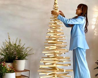 Könnte beinhalten: Eine Person schmückt einen Weihnachtsbaum aus Holz, der aus gestapelten Latten besteht. Der Baum ist hoch und schmal und hat einen Stern oben drauf. Die Person trägt einen hellblauen Blazer und eine passende Hose.