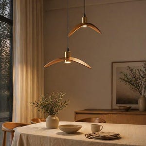 May include: Two modern wooden pendant lights with brass accents hang above a dining table. The lights have a curved, wing-like design. The table is set with a neutral-colored tablecloth, a bowl, and a vase with greenery.
