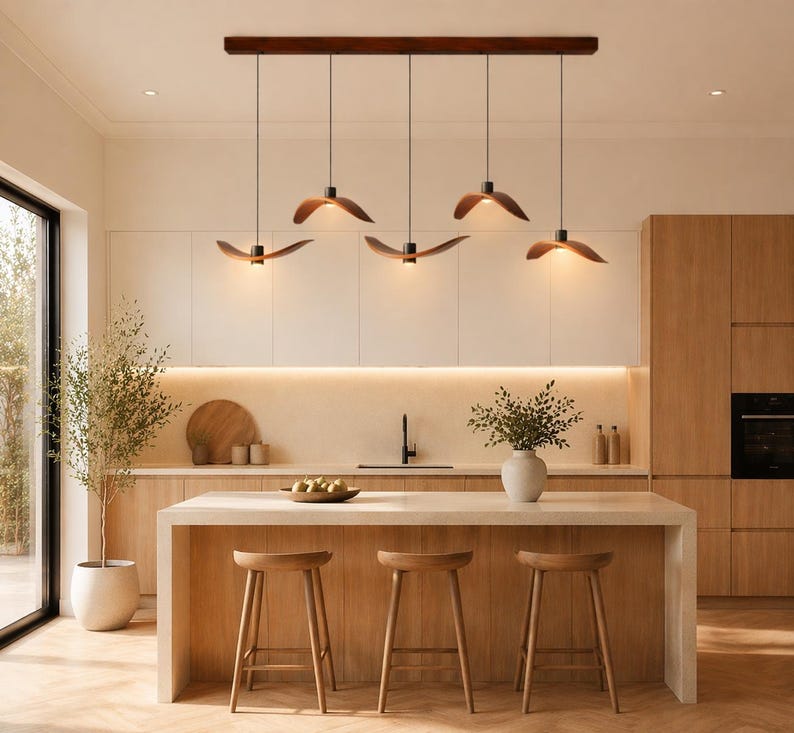 May include: A modern kitchen with a light wood island and matching stools. Above the island, five pendant lights with wooden shades hang from a dark wood beam. The kitchen features light-colored cabinets and a large window.