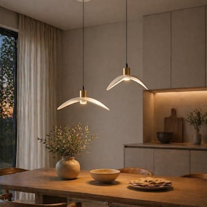 May include: Two modern pendant lights with a white, curved shade and gold accents hang above a wooden dining table. The lights are suspended from the ceiling by black cords. A vase with greenery and a wooden bowl are on the table.