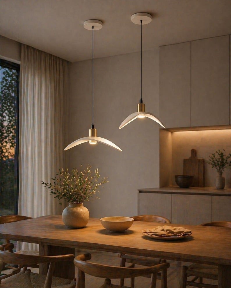 May include: Two modern pendant lights with a white, curved shade and gold accents hang above a wooden dining table. The lights are suspended from the ceiling by black cords. A vase with greenery and a wooden bowl are on the table.