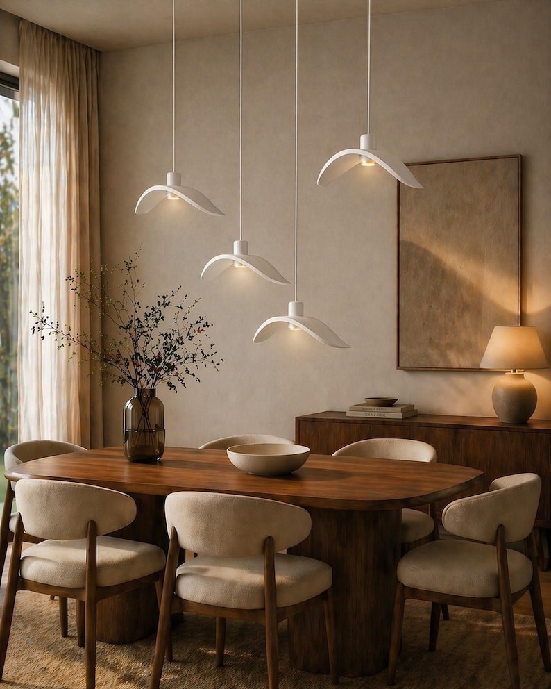 May include: A dining room scene featuring a large wooden table surrounded by six beige upholstered chairs. Above the table, five white, petal-shaped pendant lights hang from the ceiling, illuminating the space. A vase of greenery and a decorative bowl adorn the table.