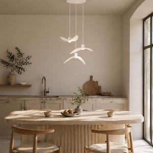 May include: A modern kitchen features a white pendant light fixture with three bird-shaped lights. The kitchen island is made of light-colored wood, with two wooden chairs. The walls are a neutral beige color, and there is a large window.