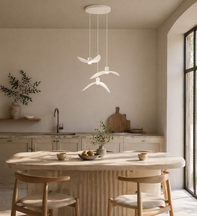 May include: A modern kitchen features a white pendant light fixture with three bird-shaped lights. The kitchen island is made of light-colored wood, with two wooden chairs. The walls are a neutral beige color, and there is a large window.