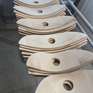 May include: Stacks of unfinished, light-colored wooden chair seats with a curved design and a central circular hole. The seats are arranged on a metal rack, suggesting a manufacturing or storage setting.