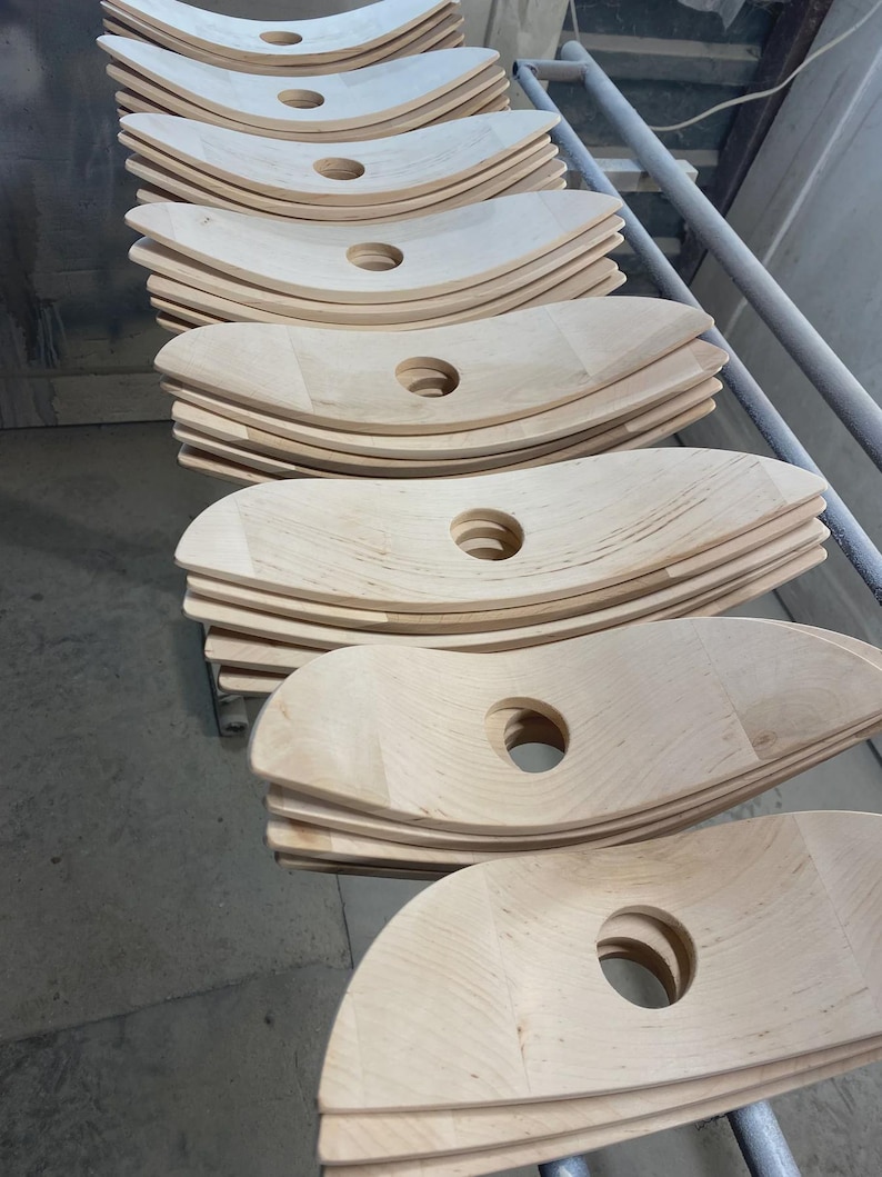 May include: Stacks of unfinished, light-colored wooden chair seats with a curved design and a central circular hole. The seats are arranged on a metal rack, suggesting a manufacturing or storage setting.