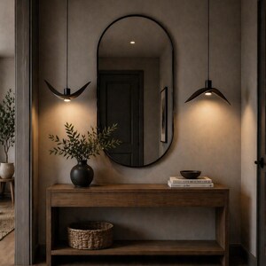 May include: A modern entryway featuring a wooden console table with a black vase holding greenery, a woven basket, and a large oval mirror. Two black pendant lights hang above. A jute rug and neutral walls complete the space.