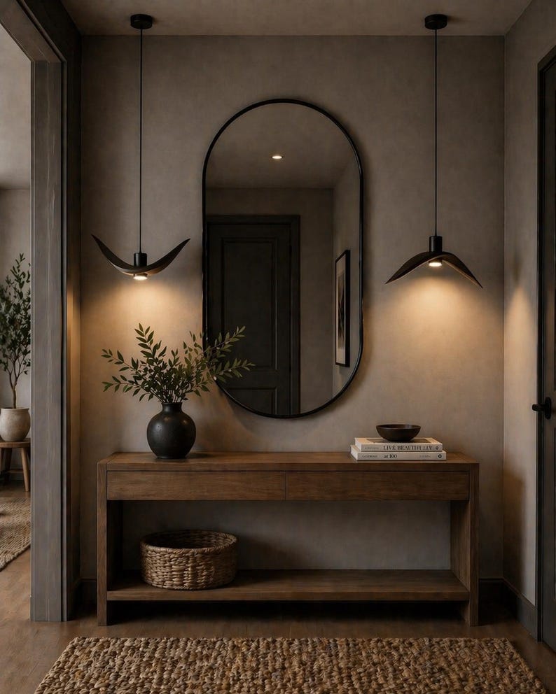 May include: A modern entryway featuring a wooden console table with a black vase holding greenery, a woven basket, and a large oval mirror. Two black pendant lights hang above. A jute rug and neutral walls complete the space.