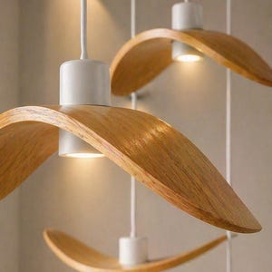 May include: Three modern pendant lights with a natural wood finish and white accents. The curved wooden shades hang from white cords, with a warm light emanating from the center of each fixture. The design is minimalist and elegant.