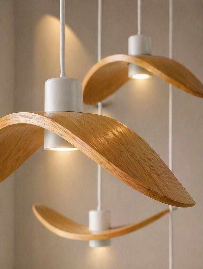 May include: Three modern pendant lights with a natural wood finish and white accents. The curved wooden shades hang from white cords, with a warm light emanating from the center of each fixture. The design is minimalist and elegant.