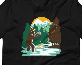 Può includere: Una maglietta nera con una grafica di un Bigfoot che indossa un cappello e pesca in un lago. La grafica è in stile retrò con una montagna e alberi sullo sfondo.