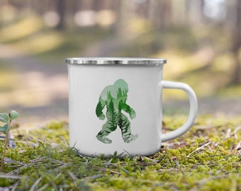 Puede incluir: Taza de camping de esmalte blanco con una silueta de Bigfoot caminando por un bosque. La silueta está llena de árboles verdes y un cielo verde y azul.