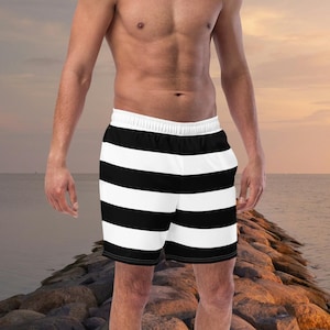 May include: Black and white striped swim shorts. The shorts have a white waistband and a black trim. The shorts are knee-length and are made of a lightweight material. The background is a beach scene with a sunset.