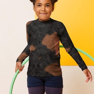 May include: A young person wearing a black and brown tie-dye long-sleeved shirt and dark blue leggings is holding a green hula hoop. They are standing on a yellow and white background.