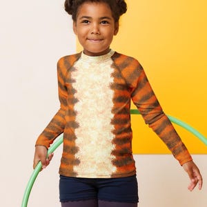 May include: A young person wearing a long-sleeved shirt with a tiger stripe pattern, navy shorts, and purple leggings, holding a green hula hoop. Pink sneakers complete the outfit. The background is a mix of yellow and off-white.