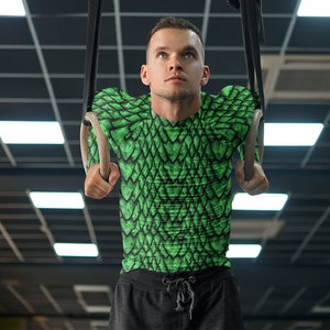 May include: A man wearing a green and black long-sleeved shirt with a scale pattern is doing a pull-up on gymnastic rings. He is wearing black shorts and has a serious expression on his face.