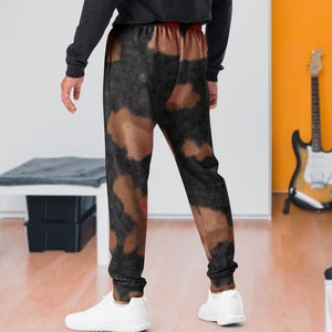 May include: A person wearing a black long-sleeved shirt and brown and black tie-dye sweatpants. The person is standing in a room with a guitar and a desk in the background.