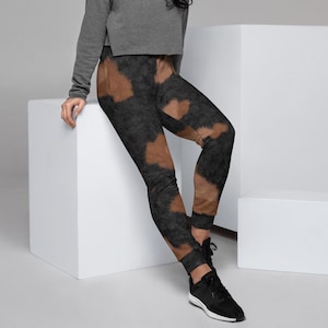 May include: A woman wearing a gray cropped sweatshirt and black and brown tie-dye sweatpants. She is standing on a white platform with her right leg bent and her left leg straight.