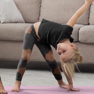 May include: Two people, a woman and a child, are doing yoga on a pink mat. They are both wearing black leggings with a brown tie-dye pattern. The woman is wearing a white tank top and the child is wearing a gray shirt. They are in a living room with a couch and a bookshelf in the background.