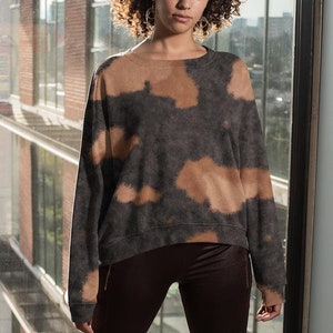 May include: A person wearing a brown and black tie-dye sweatshirt with long sleeves and dark brown leggings. They are standing in front of a large window with a city view and wearing white sneakers.