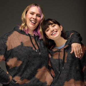 May include: Two people wearing black and brown tie-dye hoodies. The person on the left has pink hair and is smiling. The person on the right has brown hair and is smiling. They are both wearing black hoodies with brown tie-dye patterns.