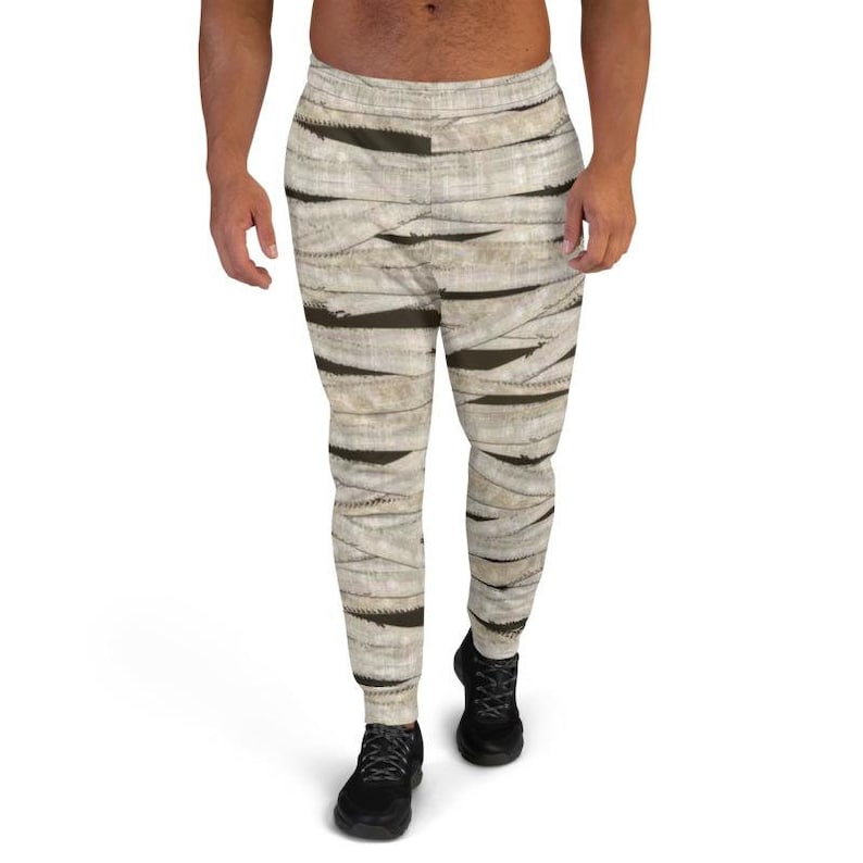 May include: A pair of sweatpants with a mummy-inspired print. The sweatpants are a light beige color with brown bandages printed on them. The sweatpants have a drawstring waistband and a relaxed fit.