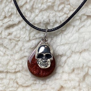 May include: A silver skull pendant necklace with a teardrop-shaped, glittery, red and brown resin base. The pendant hangs from a black cord. The necklace is displayed against a white, textured background.