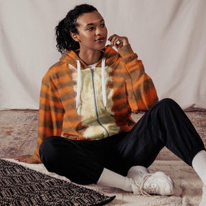 May include: A tie-dye zip-up hoodie in shades of orange and brown. The hoodie features a white drawstring and a full-length zip. The person in the image is wearing black trousers and white trainers.