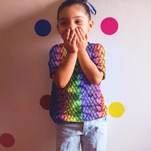 May include: A vibrant, short-sleeved top with a scale-like design in rainbow colours. The top is paired with light blue jeans. The background has colourful polka dots on a white wall.