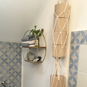 Könnte beinhalten: Badezimmer-Interieur mit einem Makramee-Toilettenpapierhalter, gefüllt mit braunen Papierrollen. Ein rundes, zweistöckiges Regal zeigt gefaltete Handtücher und Ornamente. Die Wände sind mit blau-weiß gemusterten Fliesen dekoriert.