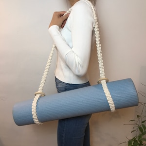 Könnte beinhalten: Ein weißer Baumwoll-Yogamatte-Gurt mit Holzringen. Der Gurt wird über die Schulter getragen und hält eine blaue Yogamatte.