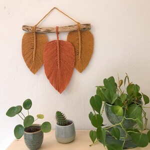 Könnte beinhalten: Ein Wandbehang mit drei Makramee-Blättern in Orange- und Gelbtönen. Die Blätter sind an einem natürlichen Ast befestigt. Der Wandbehang wird vor einer weißen Wand gezeigt.