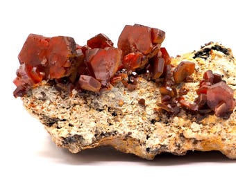 May include: A cluster of vibrant, reddish-orange crystals, likely a mineral specimen, sits atop a beige, textured matrix. The crystals have a sharp, angular structure, contrasting with the rough base. The specimen is displayed against a white background.